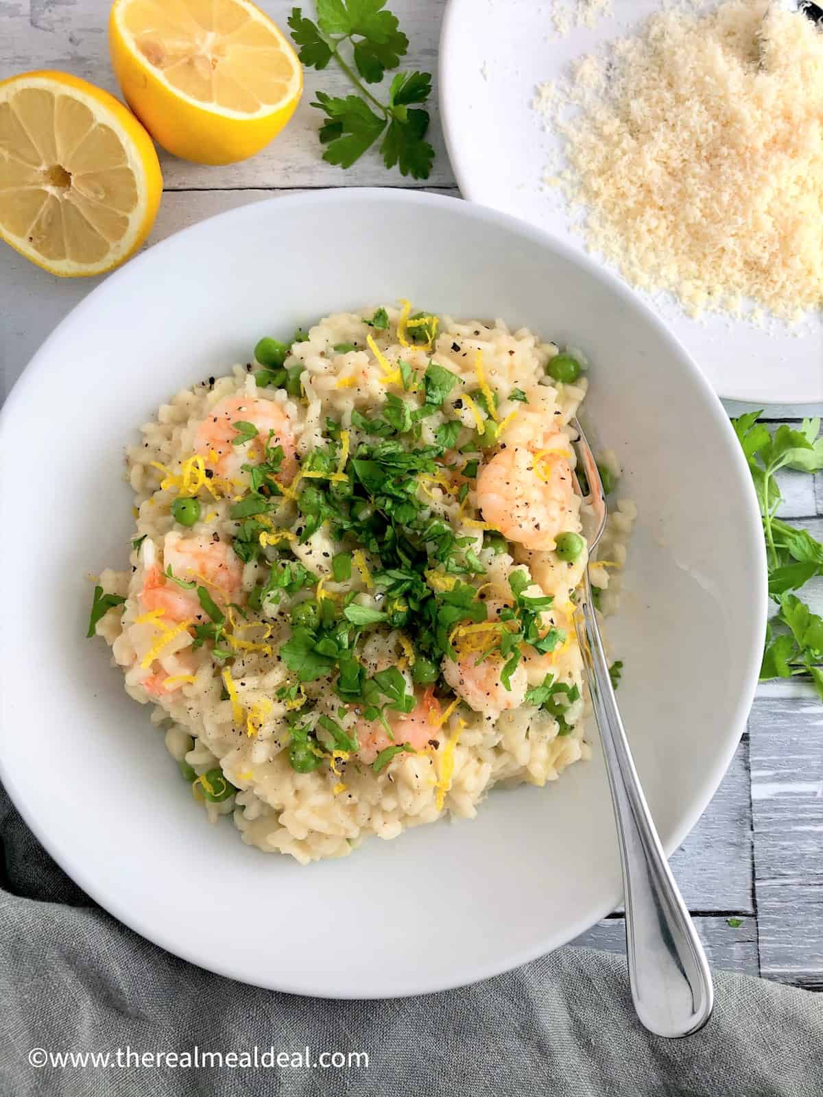 prawn and pea risotto in bowl topped with fresh parsley leaves lemon zest lemon halves parsley leaves and grated parmesan to side of dish