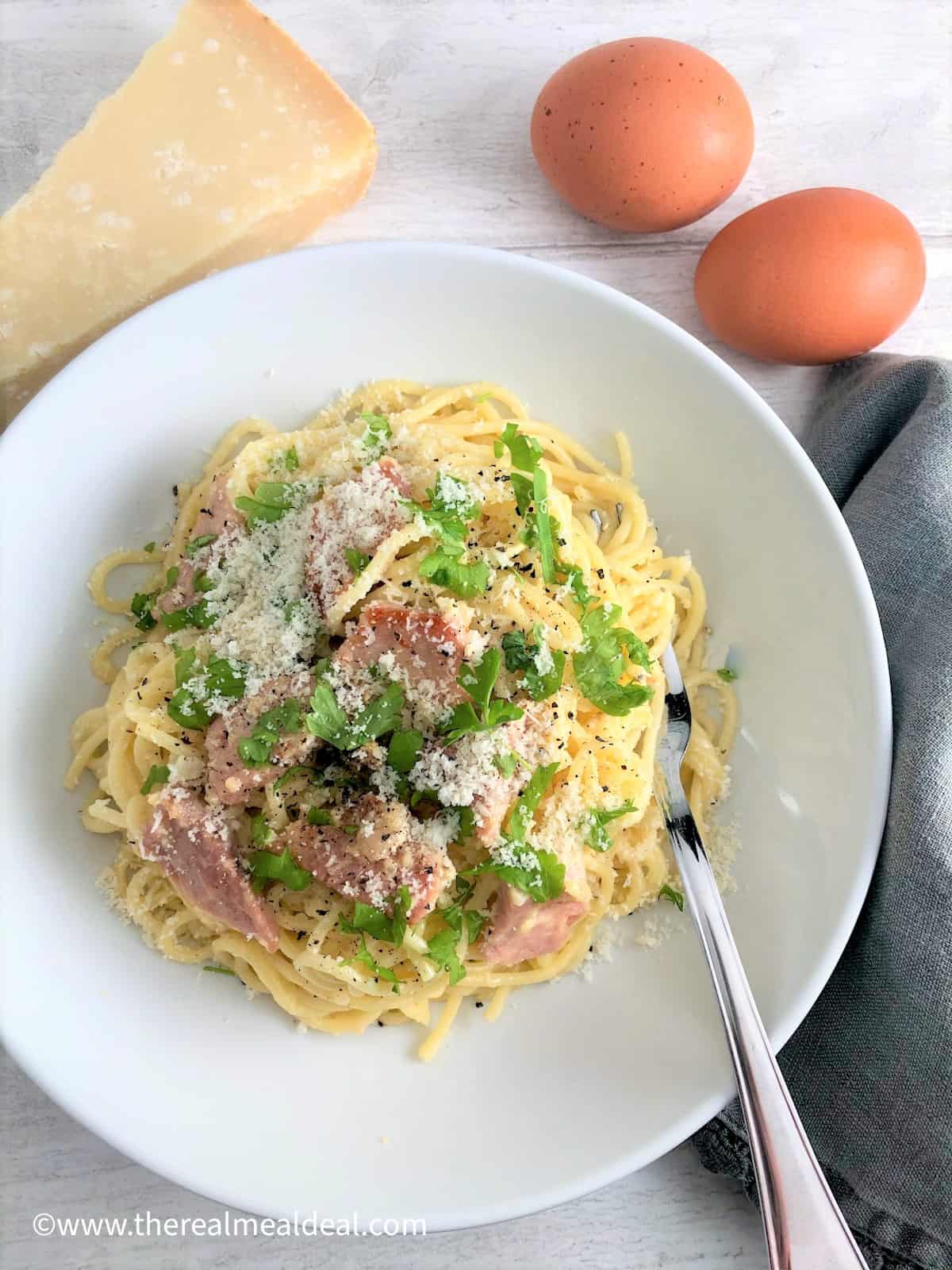 spaghetti carbonara in bowl topped with parmesan cheese parsley leaves eggs and parmesan cheese in background