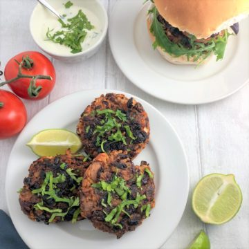3 spicy bean burgers on plate topped with fresh coriander leaves bean burger in bun in background with tomatoes and yoghurt dip