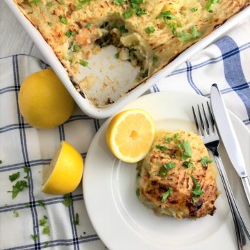 baked fish pie with leeks in dish with portion removed to plate at side served with lemon halves and fresh parsley leaves