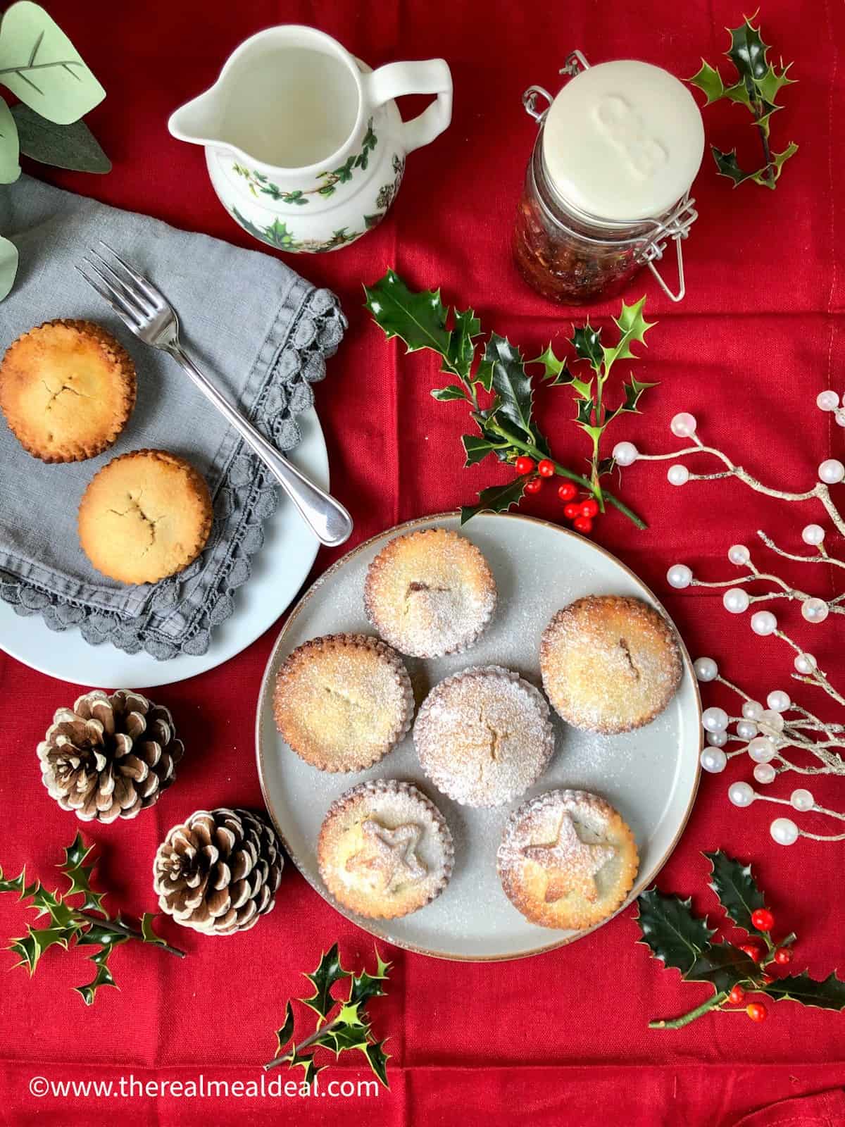Plate of Christmas mince pies dusted with icing sugar on red cloth surrounded by christmas holly decoration