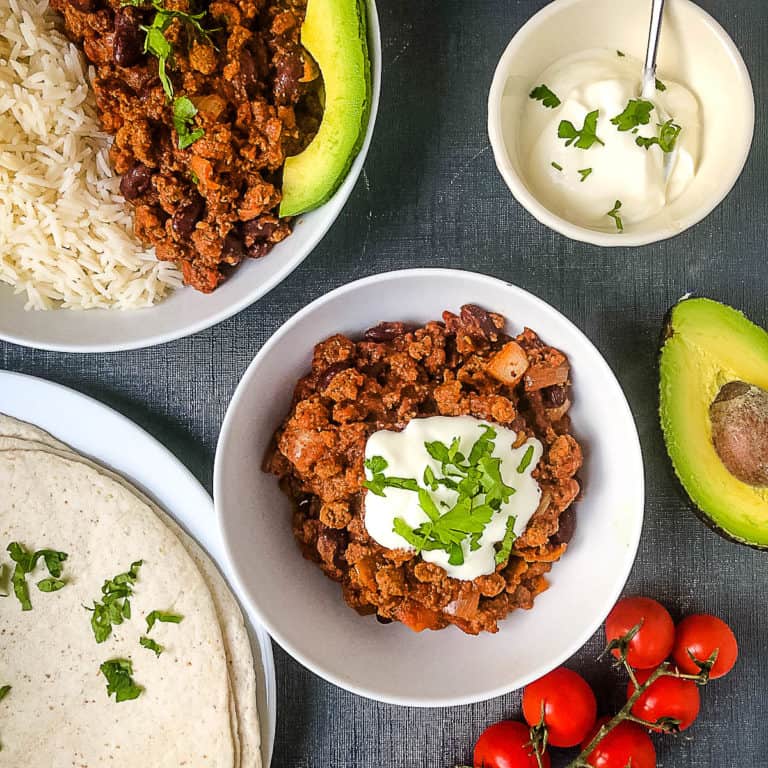 Quorn Chilli in bowl topped with soured cream, second bowl to side with chilli and rice, wraps, tomatoes, avocado and soured cream surronding bowls.
