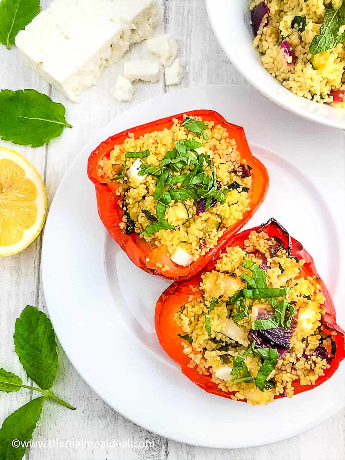 two red pepper halves stuffed with vegetable couscous and feta cheese on plate with lemon, mint and feta cheese to side